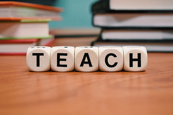 lettered blocks lined up on top of a desk spell the word teach, a stack of books and a blackboard are in the background