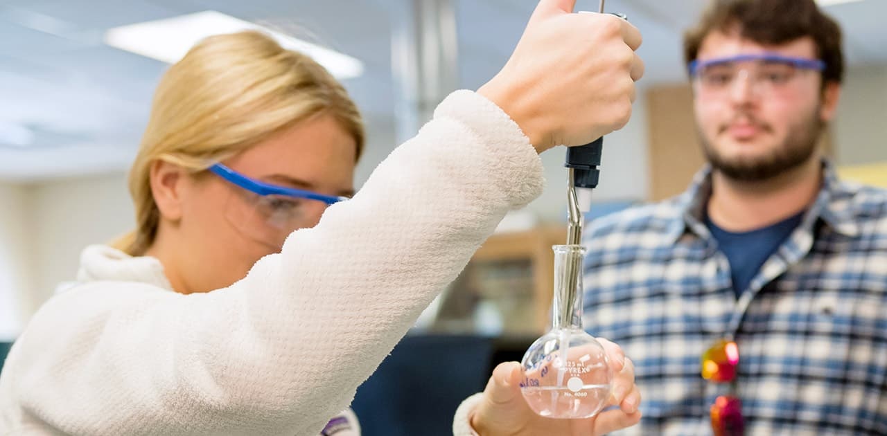 a student adds a substance to a beaker using an eyedropper while another student observes in the background