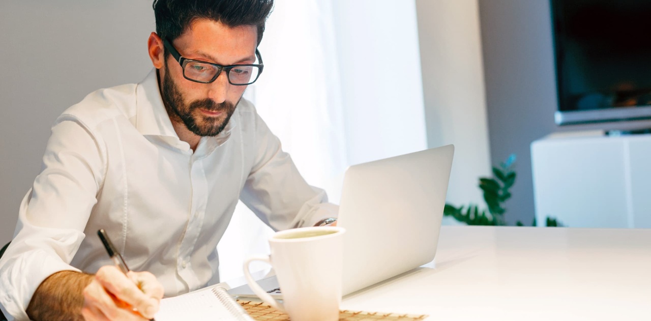 man in business shirt and classes takes notes at his kitchen table while on his laptop