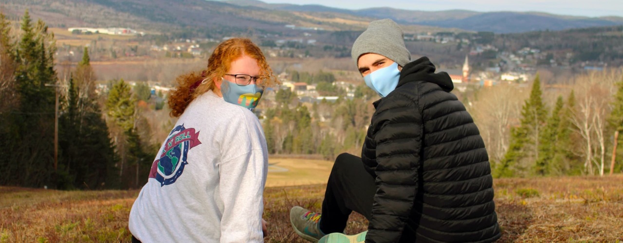 two students sit together at the top of a hill during autumn. The two students have their heads turned to the camera with a view of Fort Kent in the background.