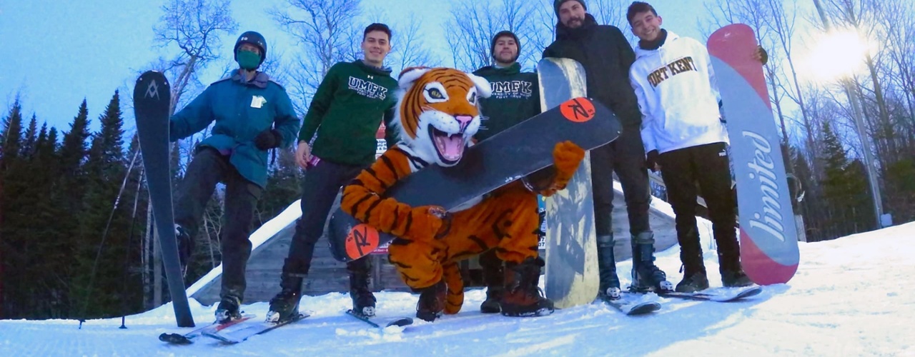 a group of students at the top of a ski hill preparing to snowboard with Benny the Bengal