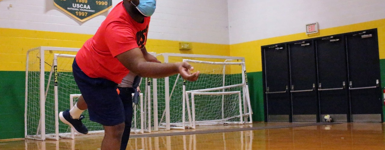 a male student, wearing a mask, tosses a bean bag while playing cornhole