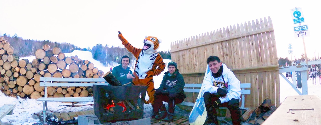 students and Benny the Bengal build a fire in an outdoor fireplace. The students sit on benches around the file while Benny stands pointing.